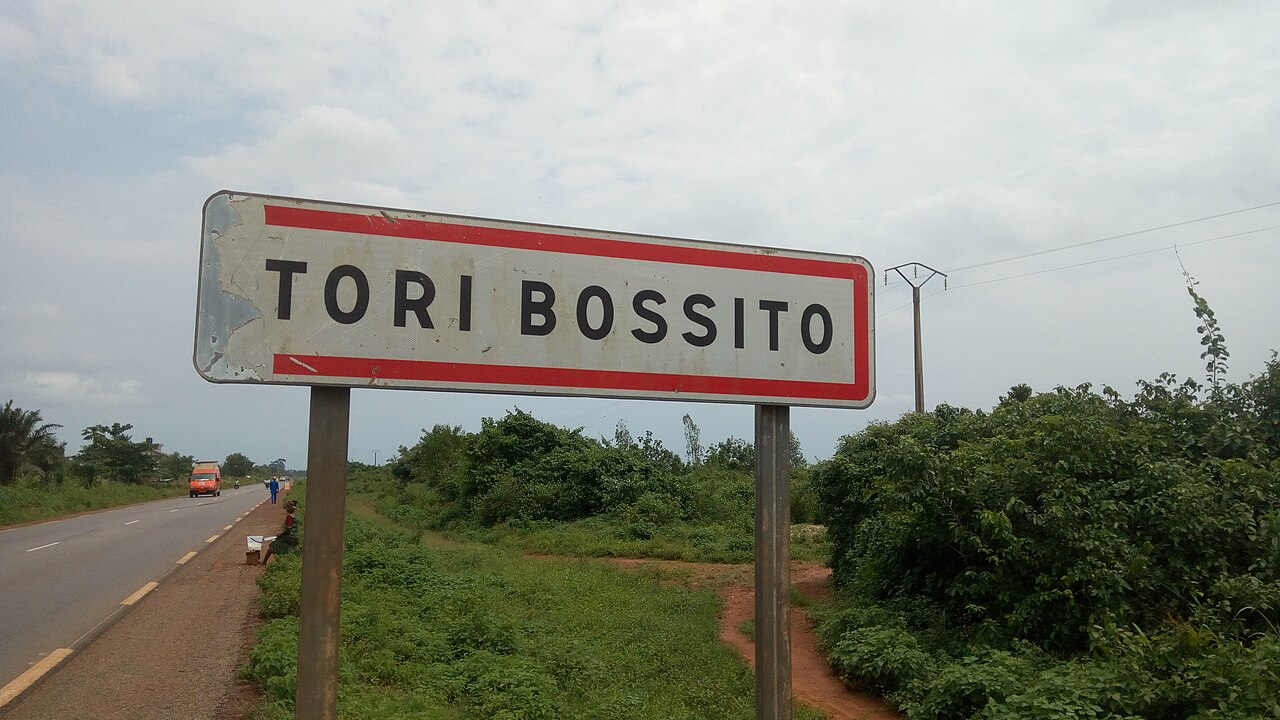 Panneau d'entrée de Tori-Bossito en bordure de route bitumée, végétation environnante